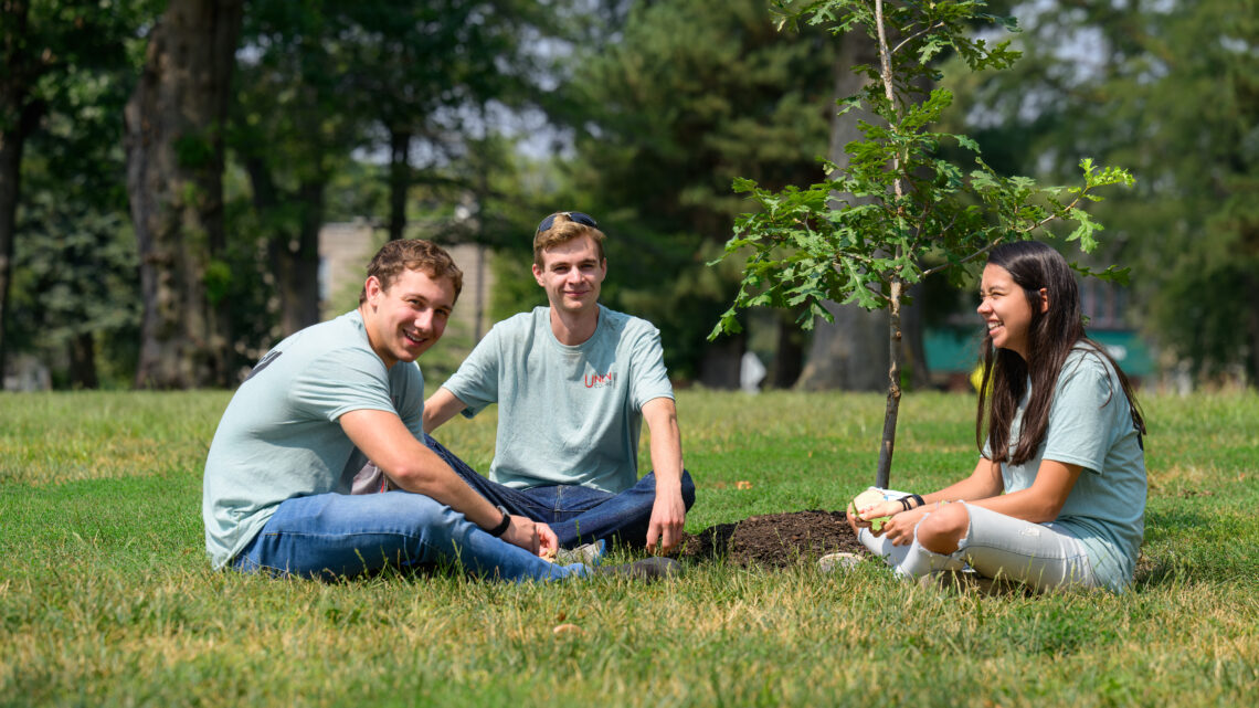 Photo of three students sitting under a sapling.